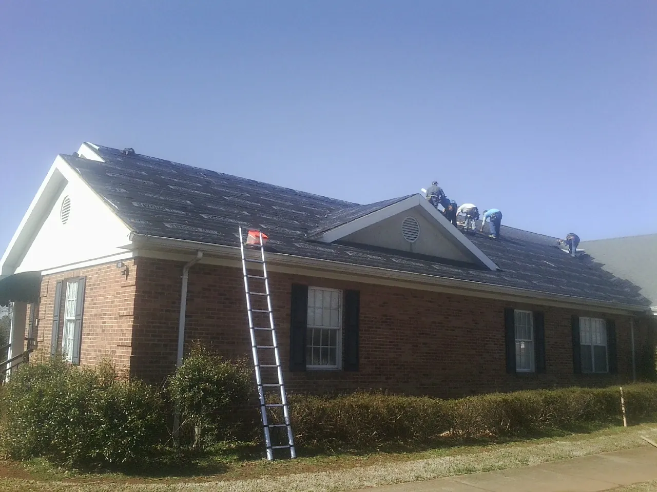 Crew tearing shingles off a brick ranch roof with a ladder set — Emergency Roof Repair work by our crew in Cana