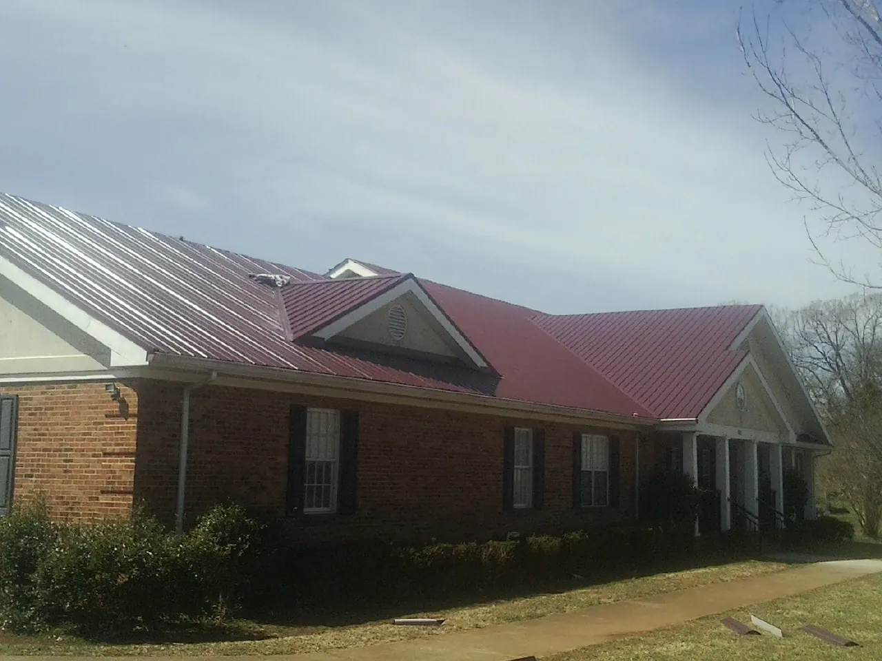 Red metal roof with gabled entries on a long brick building, from a Hail Damage Roof Repair job in Cana