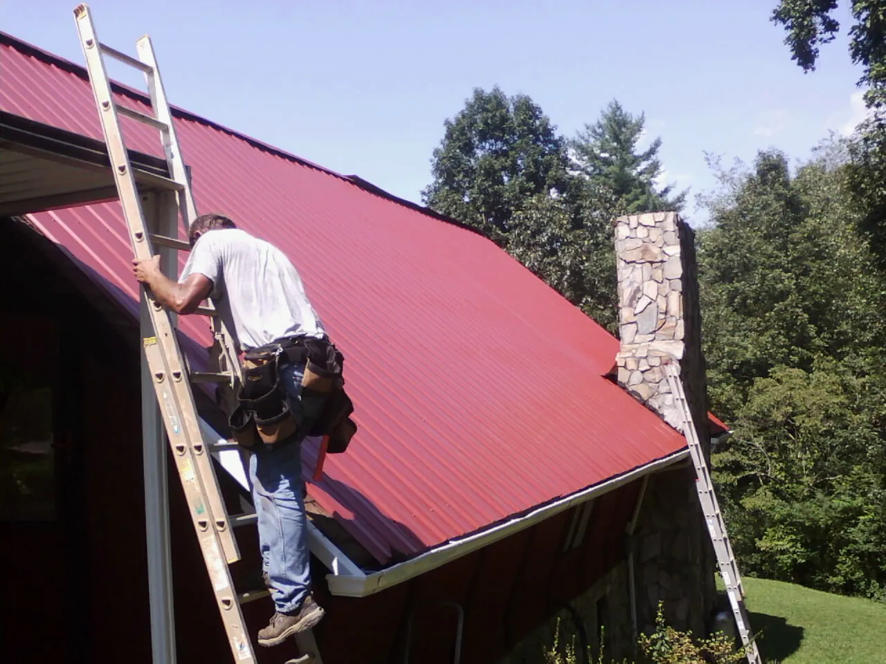 Roofer on a ladder beside a red metal roof and stone chimney — Soffit & Fascia Repair by Roofing Brothers in Cana